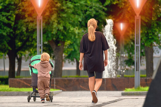 Lonely woman rests in a city park with children. A little boy is pushing a baby carriage with his sister, his mother is walking side by side.