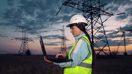 Employee with helmet looks at power transmission lines and types on laptop