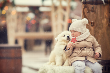 Little girl in beige winter clothes on Christmas fair eating lollipop with little white dog samoyed 