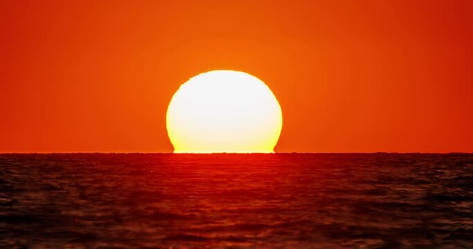 Sunset video for background. Bright orange sunset over water, huge disk of sun goes beyond horizon, as if plunging into water. Nature weather orange sky. Grey clouds. Cinematic video with no people