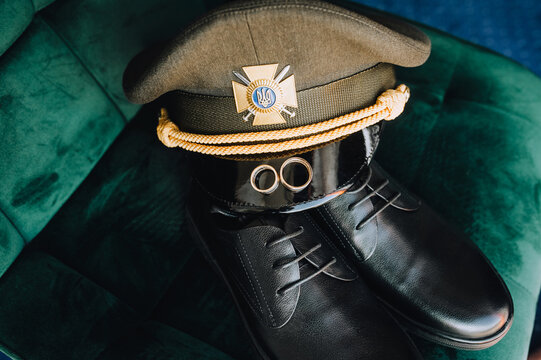 Gold rings, a cap, shoes, the uniform of a Ukrainian military groom on a chair. Wedding photography, accessories, details. War in Ukraine.