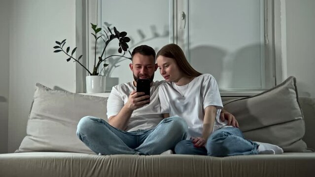 New married couple sitting at home, looking at mobile phone, scrolling browsing internet and booking tickets, hotels online for planning trip. Using gadgets and app for travelling 