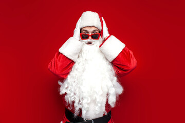 shocked santa claus in hat and festive glasses is surprised and holds his hands on his head on red background