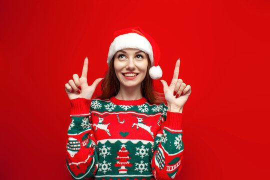 cute girl in christmas sweater and santa hat shows her hands up on red background, woman in christmas clothes