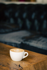 Coffee cup on wooden table on vintage sofa background. Coffee with milk in white cup. Breakfast table in vintage interior. Vertical coffee wallpaper. Latte art concept. Cappuccino in white mug.