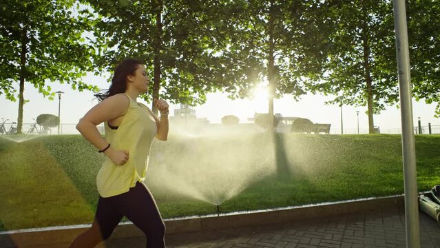 Overweight girl on a morning jog in the park. beautiful woman runs in the rays of the sun at sunrise in the city, watering the grass with water, active sports, diet, fighting with excess weight. 