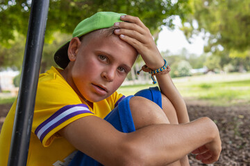 Naklejka premium a boy of European appearance in a yellow T-shirt and a green cap sits in the park with a red face and a tired look, the child plays active games in the fresh air