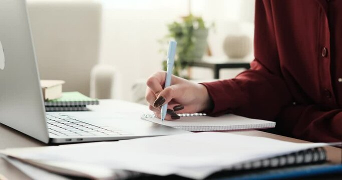 In a close-up shot, the hands of a teenage girl can be seen skillfully typing on a laptop keyboard while simultaneously making notes in a notebook using pen.