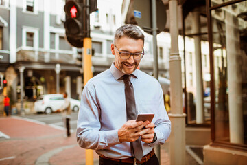 Middle aged businessman using a smartphone while walking on a sidewalk in the city commuting to work