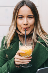 Close up of blonde young woman looking at camera while drinking orange juice on a straw. Portrait of beautiful young woman with green eyes enjoying her drink.