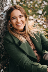 Obraz premium Close up of smiling young woman sitting and leaning on tree while looking at the camera. Portrait of natural beauty woman wearing warm clothes in autumn.
