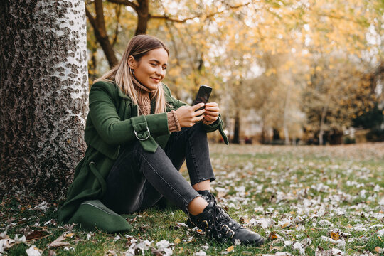 Young woman using smartphone while sitting on the ground in city park. Beautiful female sitting under a tree and texting or scrolling her social media.