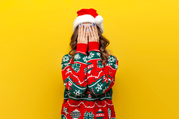 modest girl in Christmas sweater and santa claus hat covers her face with her hand and is shy on...