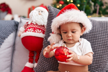 Adorable hispanic baby holding christmas decoration ball sitting on sofa at home