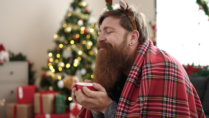 Young redhead man drinking coffee sitting on sofa by christmas tree at home