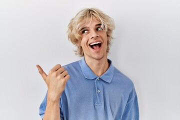 Young modern man standing over isolated background smiling with happy face looking and pointing to the side with thumb up.