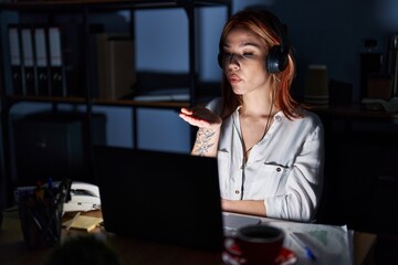 Young caucasian woman working at the office at night looking at the camera blowing a kiss with hand on air being lovely and sexy. love expression.