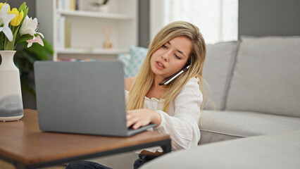 Young blonde woman using laptop talking on smartphone at home