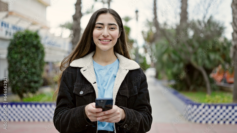 Wall mural Young beautiful hispanic woman smiling confident using smartphone at park