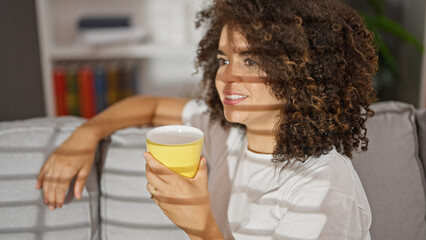 Young beautiful hispanic woman drinking coffee sitting on sofa at home