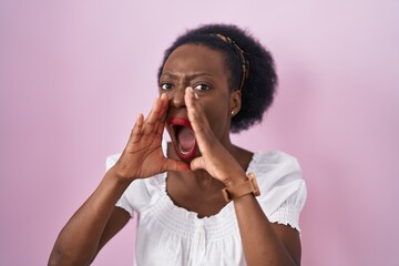 African woman with curly hair standing over pink background shouting angry out loud with hands over mouth