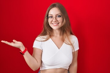 Young caucasian woman standing over red background smiling cheerful presenting and pointing with palm of hand looking at the camera.