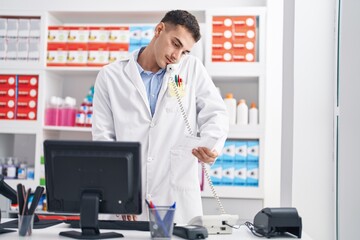 Young hispanic man pharmacist talking on telephone reading prescription at pharmacy