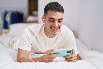 Young hispanic man watching video on smartphone lying on bed at bedroom