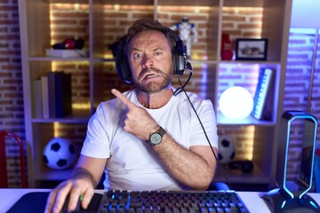 Middle age man with beard playing video games wearing headphones pointing aside worried and nervous...