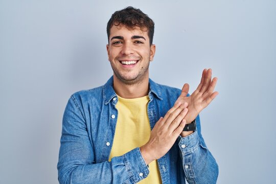 Young hispanic man standing over blue background clapping and applauding happy and joyful, smiling proud hands together