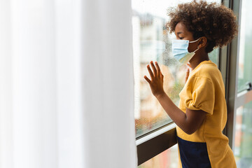 Sad lonely young girl with surgical mask looking through window while raining standing in hospital. Photo with copy space.