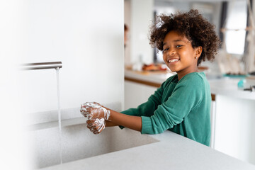 Happy young African American girl washing hands with soap smiling and looking at camera while...
