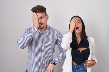 Young hispanic couple standing over white background yawning tired covering half face, eye and mouth with hand. face hurts in pain.