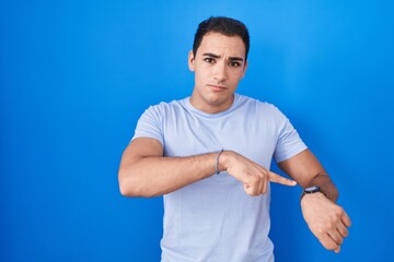Young hispanic man standing over blue background in hurry pointing to watch time, impatience, upset...
