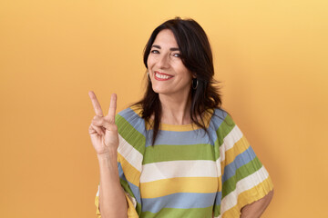 Middle age hispanic woman standing over yellow background smiling with happy face winking at the camera doing victory sign. number two.