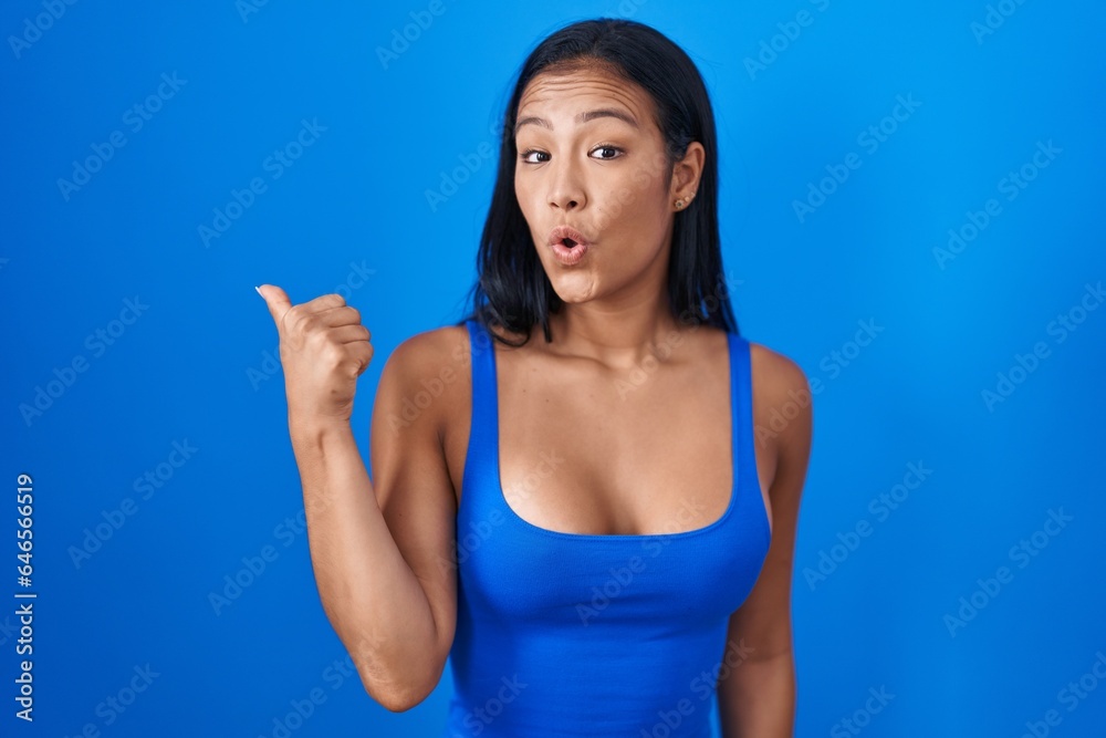 Sticker hispanic woman standing over blue background surprised pointing with hand finger to the side, open m