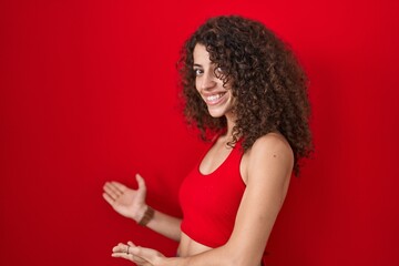 Hispanic woman with curly hair standing over red background inviting to enter smiling natural with open hand