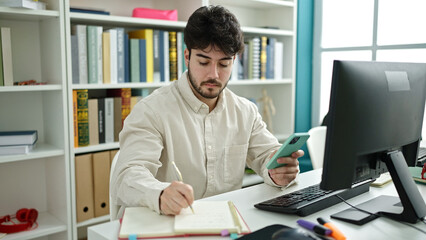 Young hispanic man student using computer and smartphone writing notes at library university
