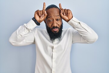 African american man standing over blue background doing funny gesture with finger over head as bull horns