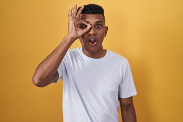 Fototapeta na wymiar Young hispanic man standing over yellow background doing ok gesture shocked with surprised face, eye looking through fingers. unbelieving expression.