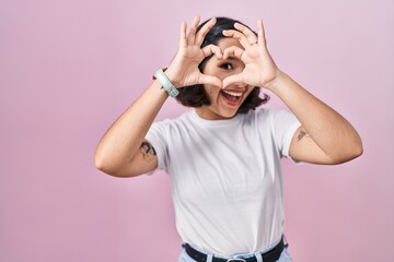 Young hispanic woman wearing casual white t shirt over pink background doing heart shape with hand...