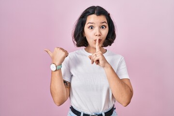 Young hispanic woman wearing casual white t shirt over pink background asking to be quiet with finger on lips pointing with hand to the side. silence and secret concept. - obrazy, fototapety, plakaty