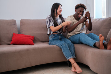 Obraz premium Young couple of different origins on the couch at home deciding what to watch on TV. Concept: lifestyle, life as a couple, bond.