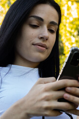 Close-up of a young woman with long dark hair holding a smartphone in her hands and browsing the news feed. Close-up of a woman's hand using a mobile phone outdoors. Girl in a white T-shirt