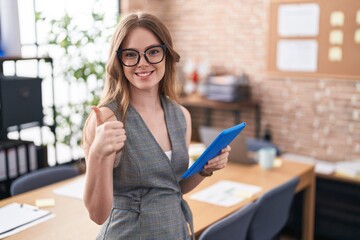 Caucasian woman working at the office wearing glasses doing happy thumbs up gesture with hand. approving expression looking at the camera showing success.