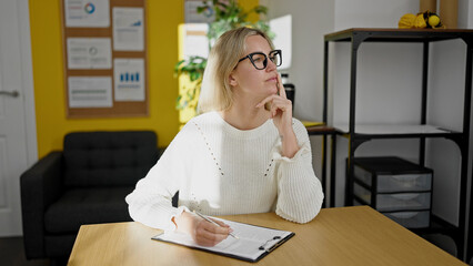 Young blonde woman business worker writing on clipboard with doubt expression at office