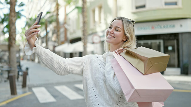 Young blonde woman going shopping holding bags making selfie by smartphone at street