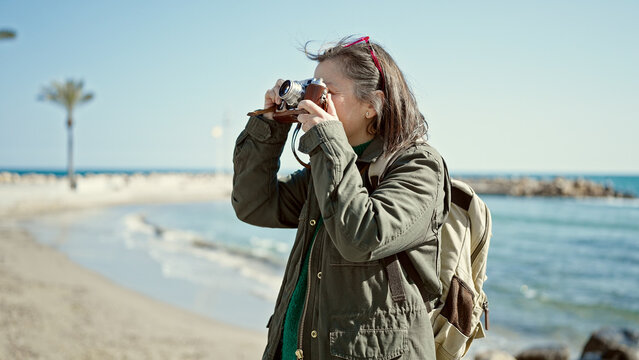 Mature hispanic woman with grey hair tourist wearing backpack taking pictures with vintage camera at seaside