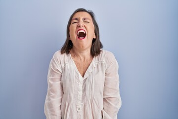 Obraz na płótnie Canvas Middle age hispanic woman standing over blue background angry and mad screaming frustrated and furious, shouting with anger. rage and aggressive concept.