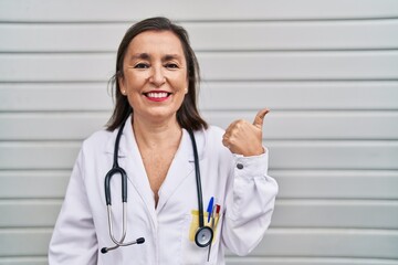 Middle age hispanic woman wearing doctor uniform and stethoscope pointing thumb up to the side smiling happy with open mouth